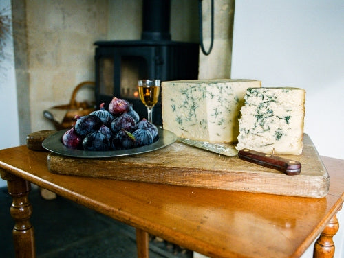 A quarter Colston Bassett Stilton sits on a cheeseboard next to a glass of Sherry and some plums. 