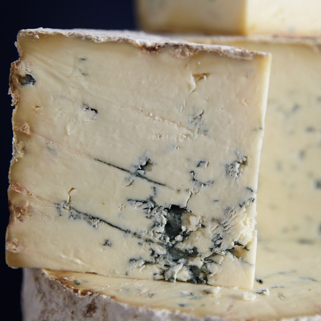 Close up of a piece of Stichelton cheese, sat on a larger cut piece of cheese, showing the developed rind, and blue veined, soft creamy paste